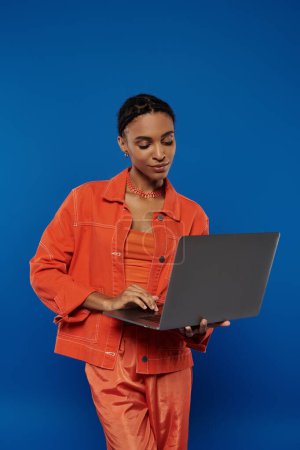 A young African American woman in an orange jumpsuit using a laptop against a blue backdrop.