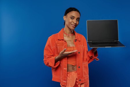 Une jeune femme afro-américaine dynamique dans une chemise orange se concentre sur la tenue d'un ordinateur portable sur un fond bleu.