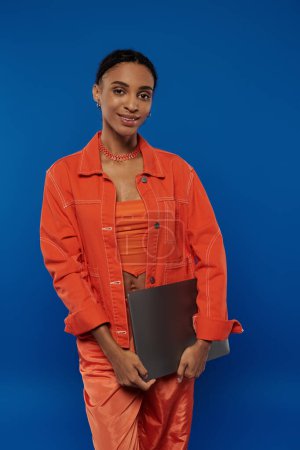 Foto de Una mujer afroamericana muy joven con un traje naranja vibrante se levanta con confianza sobre un fondo azul brillante. - Imagen libre de derechos