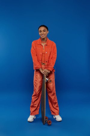A young African American woman in an orange jumpsuit holding a skateboard on a vibrant blue background.