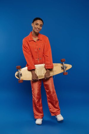 Young African American woman in orange jumpsuit confidently holds skateboard against a colorful background.