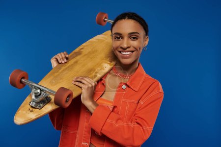 A smiling young African American woman in a vibrant orange outfit holds a skateboard against a blue background.