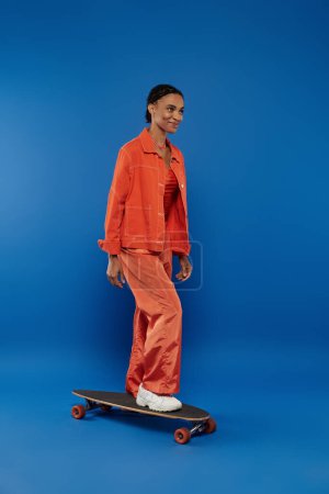A young woman in a bright orange outfit stands confidently on a skateboard against a blue background.