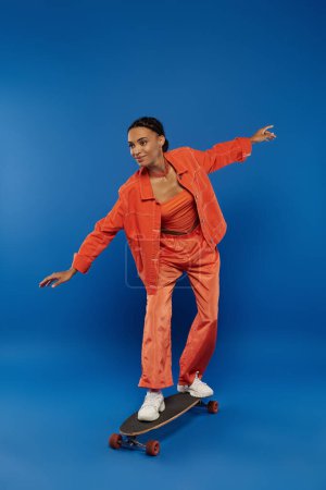 Young African American woman in orange jumpsuit skateboarding on a blue background.