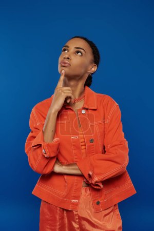 A pretty young African American woman in a vibrant orange jacket gazes upwards against a blue background.