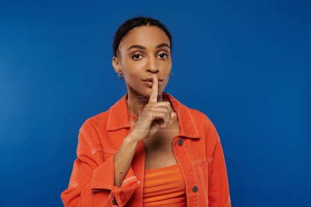Eine hübsche junge Afroamerikanerin in einem leuchtend orangefarbenen Hemd hält ihren Finger an ihre Lippen.