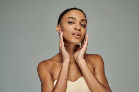 A beautiful young African American woman in a strapless top, gently touching her face with both hands, on a grey background.