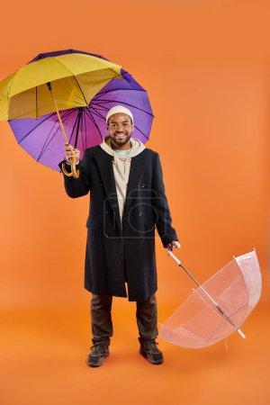 A stylish African American man in a black coat holds two umbrellas on a vibrant backdrop.
