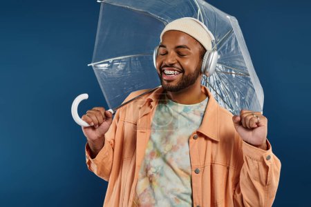 Stilvoller afroamerikanischer Mann mit klarem Regenschirm über dem Kopf vor lebendiger Kulisse.