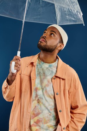 Stylish man holds clear umbrella on blue backdrop.
