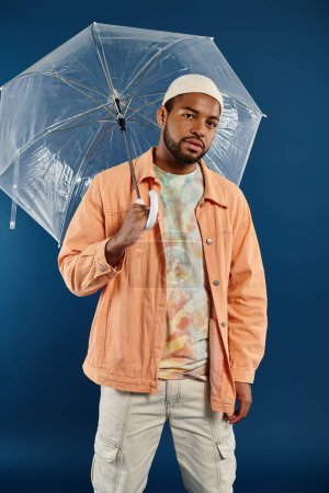 A stylish African American man holding a clear umbrella over his head against a vibrant backdrop.