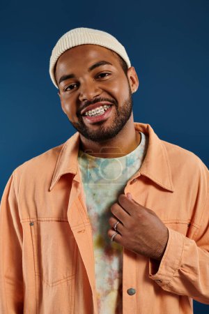 A stylish African American man posing on a vibrant backdrop wearing an orange shirt and a white hat.