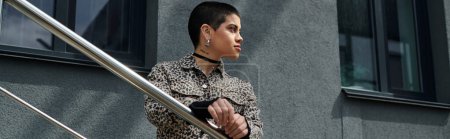 A stylish young woman with short hair and a leopard print jacket stands on a staircase next to a modern city building.