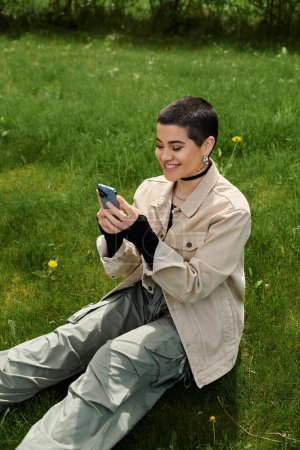 A young woman with short hair sits on lush green grass, completely absorbed in her cell phone screen.