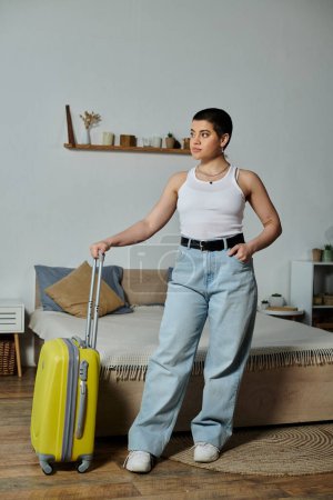 Junge Frau mit kurzen Haaren bereitet sich auf Urlaub vor, steht mit gelbem Koffer in einem Zimmer.