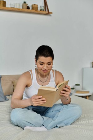 Foto de Una mujer joven con el pelo corto está sentada en una cama, absorta en la lectura de un libro en su ropa casual en casa. - Imagen libre de derechos