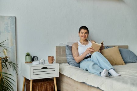 Foto de Una joven con el pelo corto, vestida casualmente, absorta en la lectura de un libro mientras está sentada en una cama. - Imagen libre de derechos