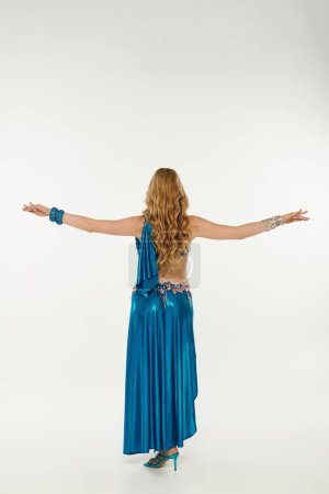 Young woman in a blue dress with arms outstretched while performing a belly dance.