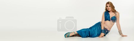 Graceful woman in blue dress resting on ground after belly dance performance.