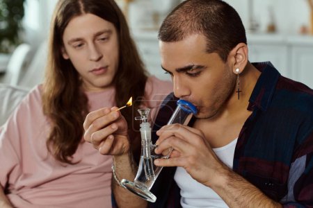 A man in casual clothes lights a bong while a partner, part of an LGBT couple, looks on in a cozy home setting.