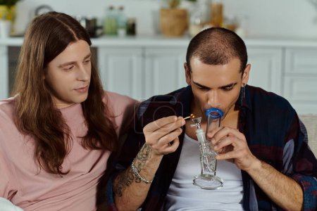 A gay couple in casual attire relaxing on a couch at home, man lighting marijuana in the glass bong