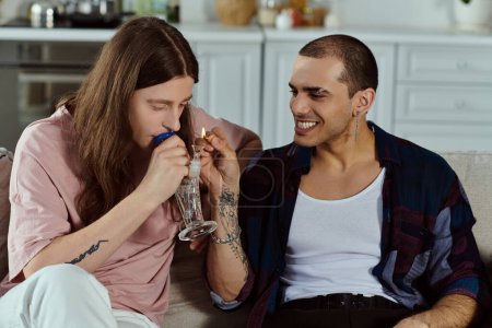 A gay couple in casual attire sit closely together on a couch, lighting marijuana in the glass bong