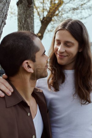 A gay couple in casual attire stands joyfully together, surrounded by lush greenery, next to a towering tree.
