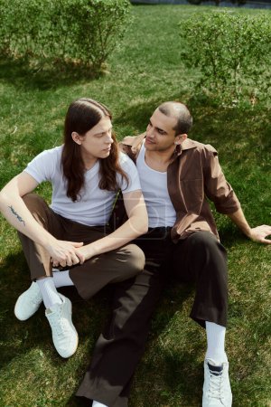 Téléchargez les photos : Gay couple dans casual tenue assis sur l 'herbe profiter un paisible moment ensemble à l' extérieur. - en image libre de droit