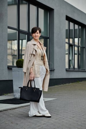 Une belle femme mature en pantalon blanc et un trench coat sur une promenade.