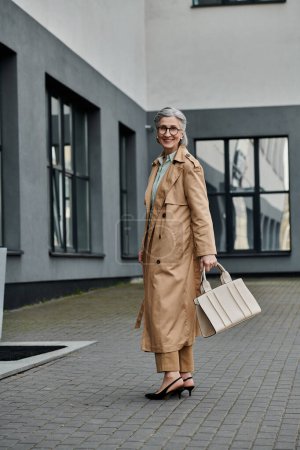 Una mujer de moda en una gabardina llevando una bolsa mientras camina con confianza.