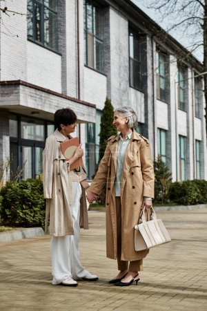 Two women, a mature beautiful lesbian couple, standing hand in hand in front of a building.