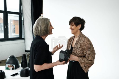 Middle-aged lesbian couple in a photo studio - one with a camera, the other modeling - engage in conversation.