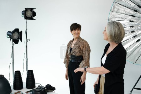 Couple lesbien d'âge moyen dans un studio ; une femme se tient devant la caméra, l'autre capture sa beauté.