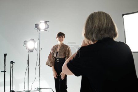 A middle-aged woman stands confidently in front of a camera in a bright, white studio.