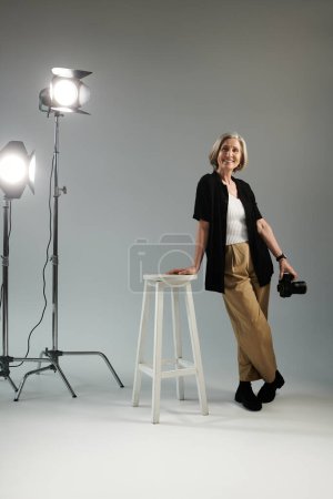 Una mujer de mediana edad está de pie junto a un taburete frente a una luz brillante del estudio, posando con la cámara