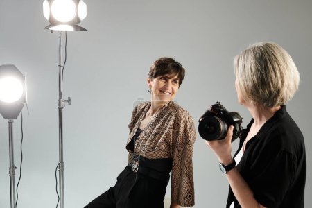 Middle aged lesbian couple in a photo studio. One woman, the photographer, is taking a picture of the other, the model.
