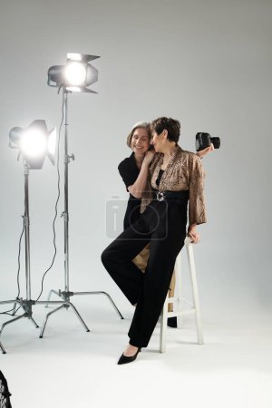 Lesbisches Paar mittleren Alters, die eine als Fotografin mit Kamera, die andere als Model, sitzen auf Hockern im Fotostudio.