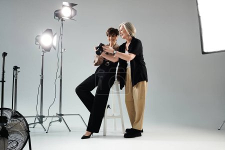 A middle-aged lesbian couple in a photo studio; one poses as the model while the other showing photos with a camera.