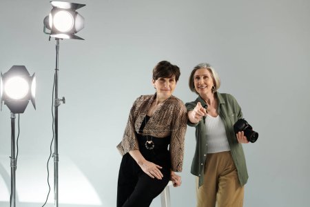 Ein lesbisches Paar mittleren Alters in einem Fotostudio, das eine mit der Kamera festhält, das andere als Model posiert.