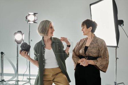 Ein lesbisches Paar mittleren Alters in einem Fotostudio; eine Frau, eine Fotografin, hält eine Kamera, während sie dem Model Posen erklärt.