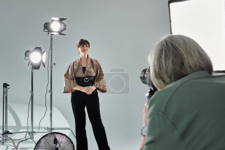 Middle-aged lesbian couple in a photo studio, one taking photos with a camera, the other posing as the model.