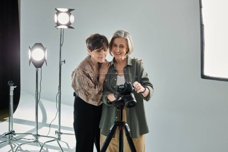 A middle-aged lesbian couple in a photo studio, one with a camera and the other as a model, standing next to each other.