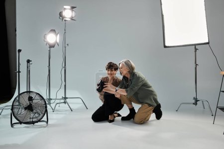 A photographer and model, a middle aged lesbian couple, kneel before the camera in a photo studio.