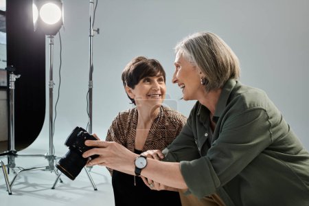 A middle-aged lesbian couple in a photo studio; one woman using a camera, and other smile
