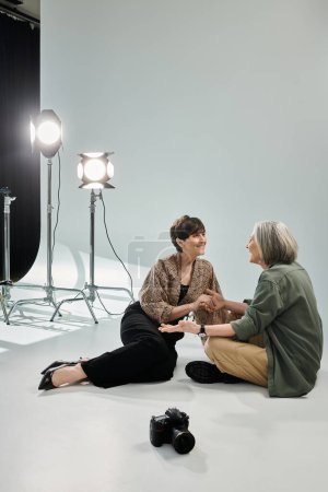 A middle-aged lesbian couple, one as a photographer with a camera, the other as a model, sitting in a photo studio.