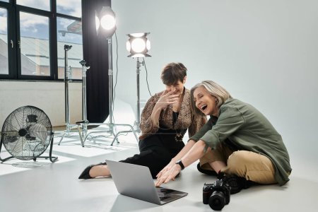 A middle-aged lesbian couple sits on the floor, engrossed in a laptop, brainstorming and creating content together.