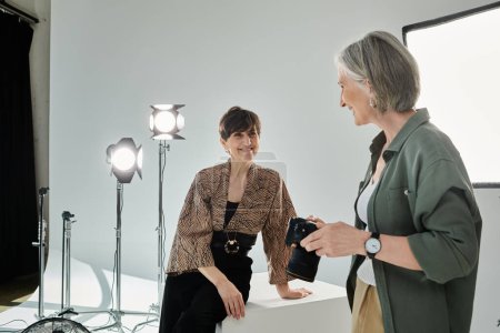 Photo for A photographer and model, a middle-aged lesbian couple, stand side by side in a photo studio, one holding a camera. - Royalty Free Image