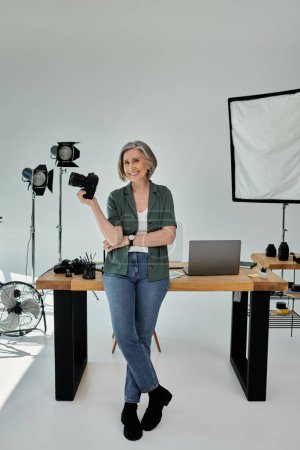 Una mujer de mediana edad se para en un estudio fotográfico, sosteniendo una cámara frente a un escritorio.
