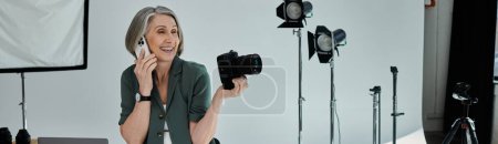 A middle-aged woman holding camera and talking on smartphone in a vibrant photo studio with professional equipment and lighting.