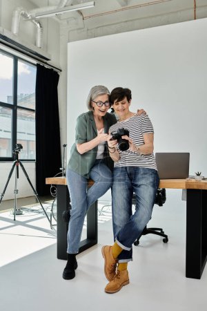 A middle aged lesbian couple collaborates on a project while sitting together on a table in a modern studio.